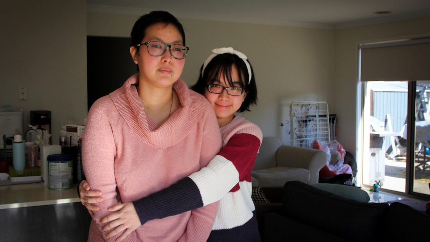 A taller woman in her 30s stands in a kitchen lounge with a younger woman hugging her from behind