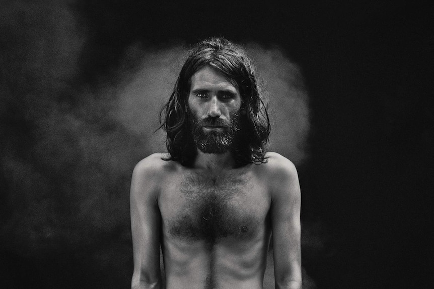 Black and white portrait photograph of Behrouz Boochani standing shirtless in front of a black background and small fire flames.