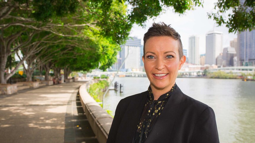 A woman with short, dark hair, wearing a black jacket, standing near a walkway next to the Brisbane River.