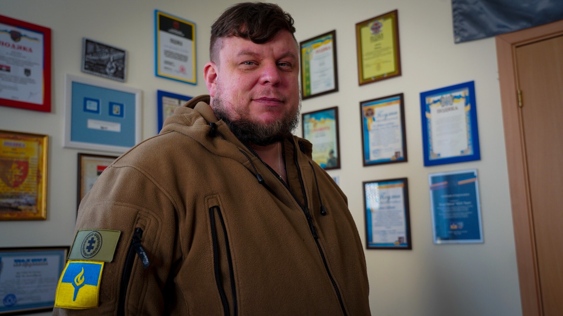 A bearded man in a brown jacket looks at the camera softly.