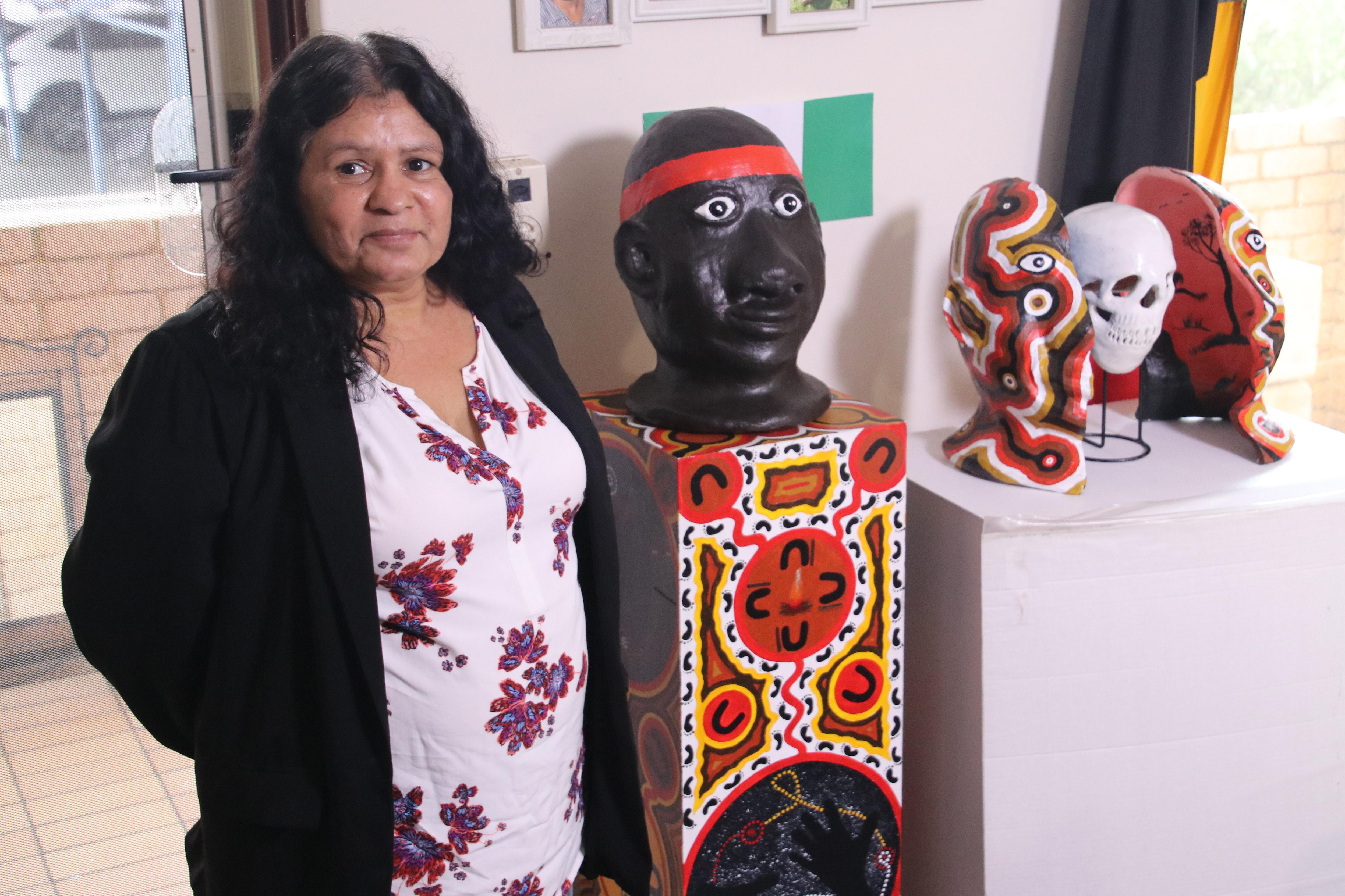 An Indigenous woman stands smiling for a photo next to a sculpture of a head and other artworks.