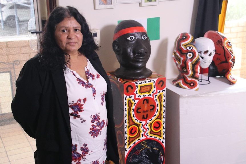 An Indigenous woman stands smiling for a photo next to a sculpture of a head and other artworks.