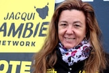 A middle-aged woman stands in front of a yellow sign which reads: Jacqui Lambie Network Vote 1