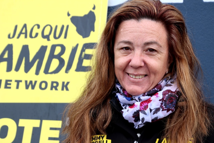 A middle-aged woman stands in front of a yellow sign which reads: Jacqui Lambie Network Vote 1