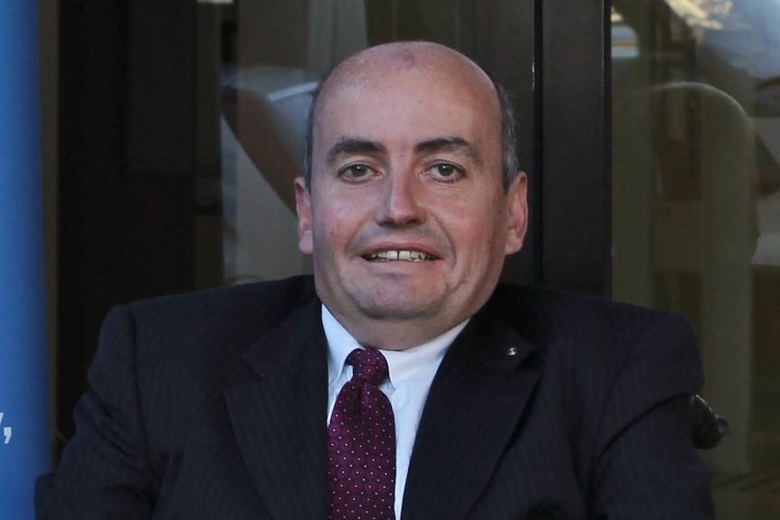 A man with a bald head, maroon tie and black suit with a white shirt