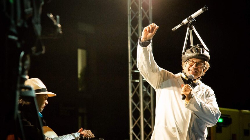 Charley Lineweaver wears a bike helmet with a telescope on top while speaking into a microphone on stage.