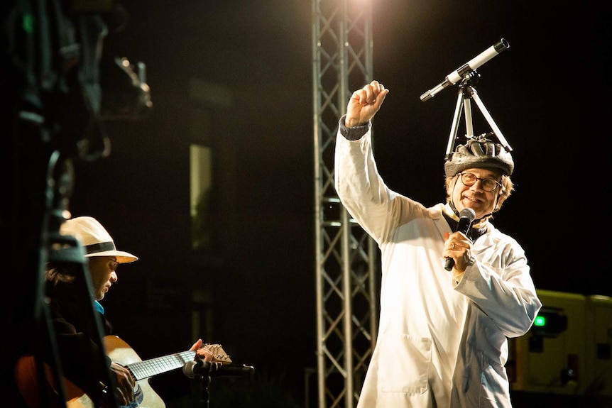 Charley Lineweaver wears a bike helmet with a telescope on top while speaking into a microphone on stage.