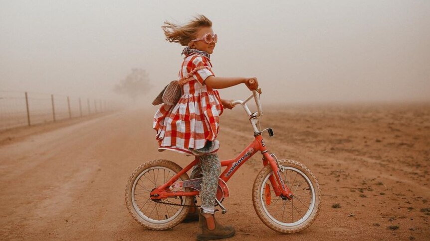 Small girl on a pink bike wearing goggles stand defiantly in dust storm