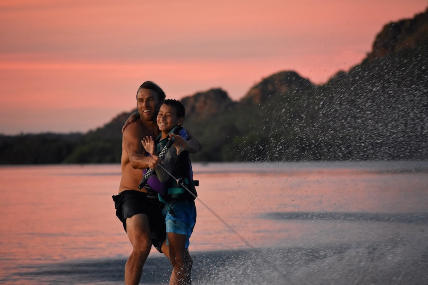 Man and young boy, his son, on surfboard being towed behind boat, with hills in background.