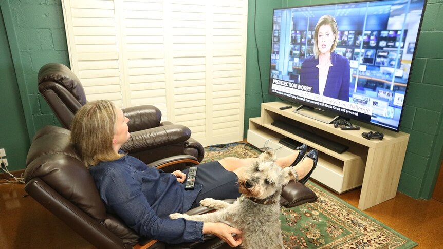 A woman sits in an armchair watching tv next to a dog