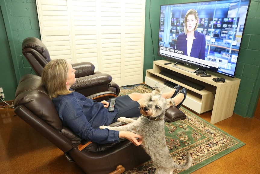 A woman sits in an armchair watching tv next to a dog