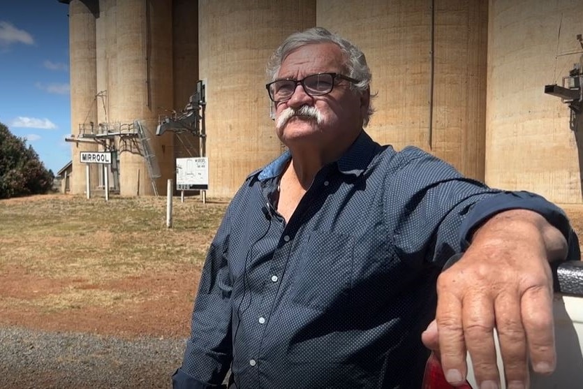 A man with a moustache and black glasses sits in front of grain silos.