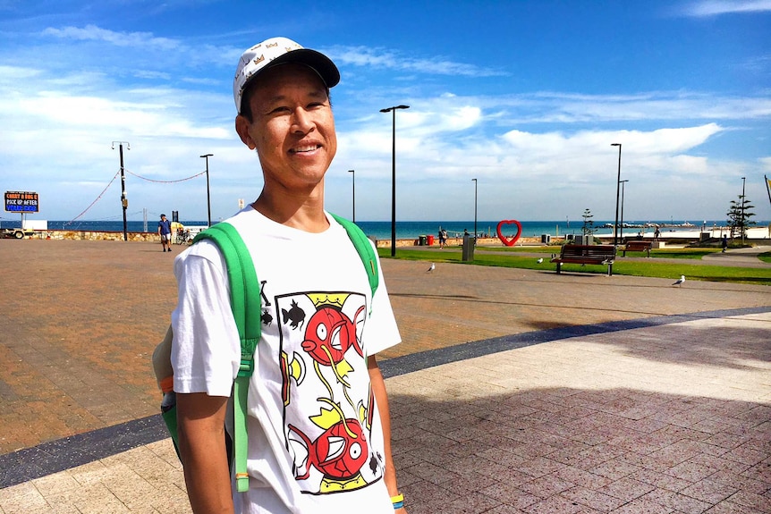 Kym, wearing a cap and t-shirt, stands in front of a coastal backdrop
