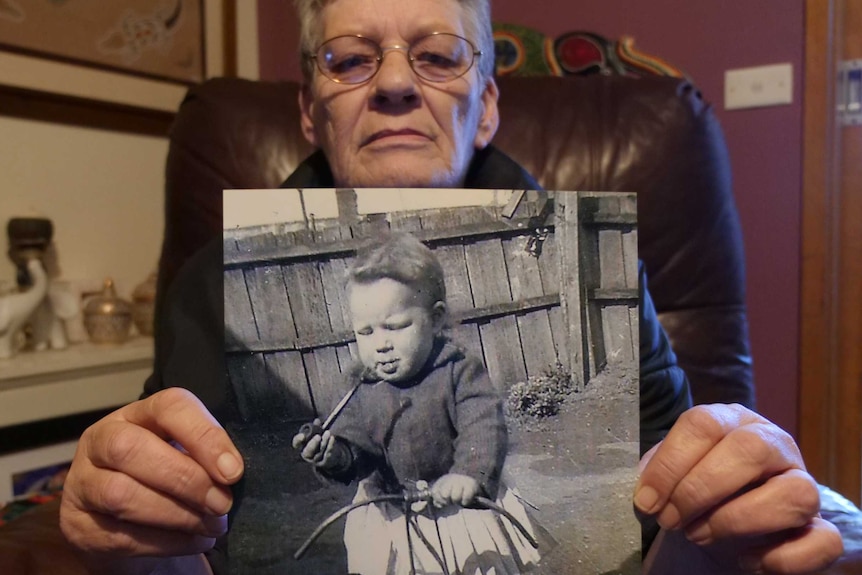 An old woman wearing glasses holding a black and white photo of herself when very young.