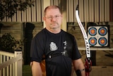 A man with an archery bow standing at a short distance from a target in his backyard.