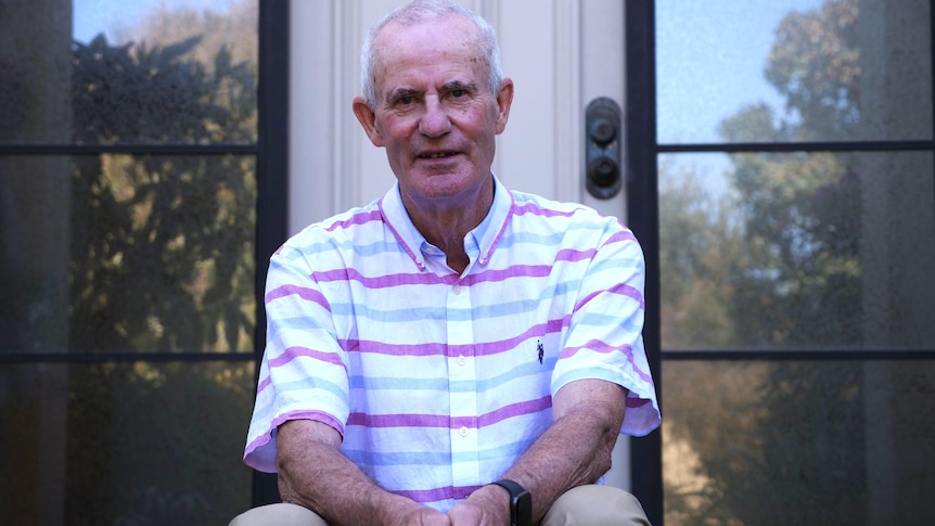 An older  man sits on a steps at a house wearing a pink striped shirt