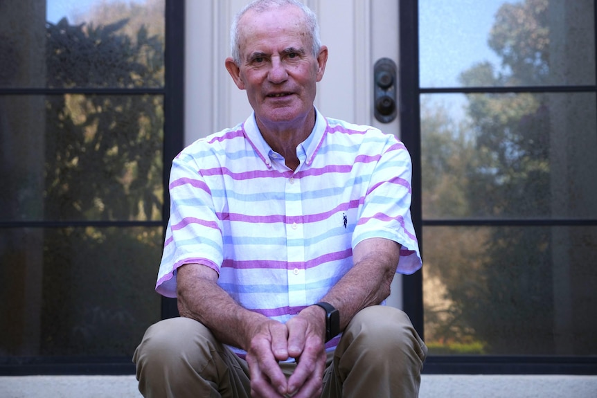 An older  man sits on a steps at a house wearing a pink striped shirt