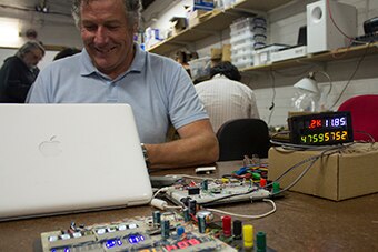 A man sits behind a laptop, surrounded by wires and alpha-numeric LED displays