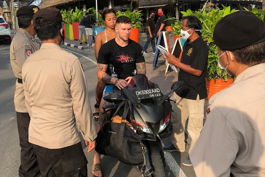 A couple on motorbike without wearing helmet and masks and surrounded by police officers.