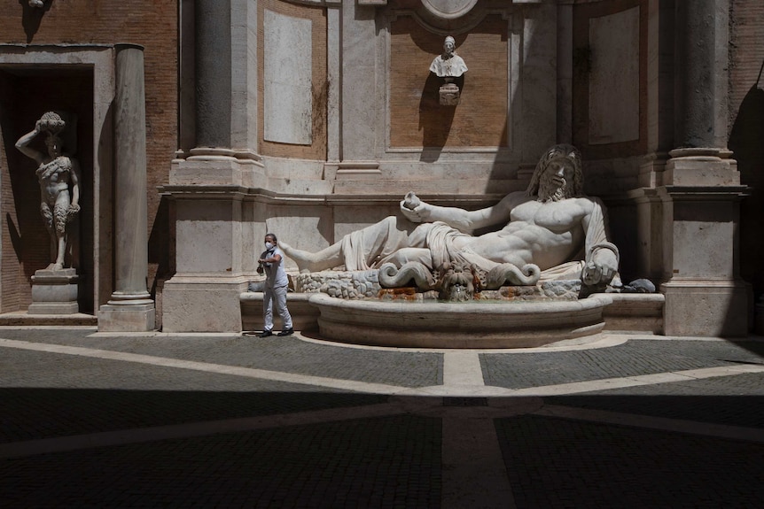 A person wearing a face mask fastens the lid of a bottle while walking from a marble fountain in which a bearded deity reclines.