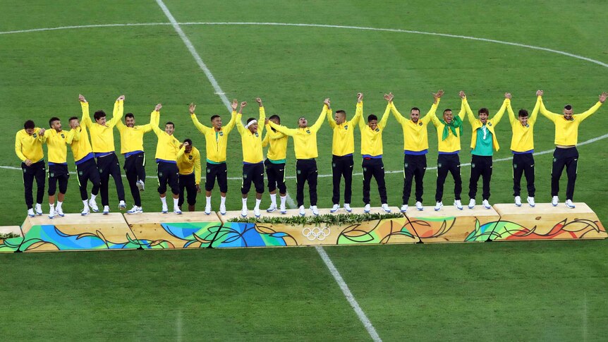 The Brazilian football team, line up on a podium, throw their hands in the air in celebration.