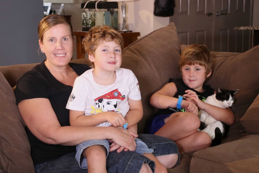 A mother sits on a brown sofa at home smiling with her two sons, aged 6 and 7.