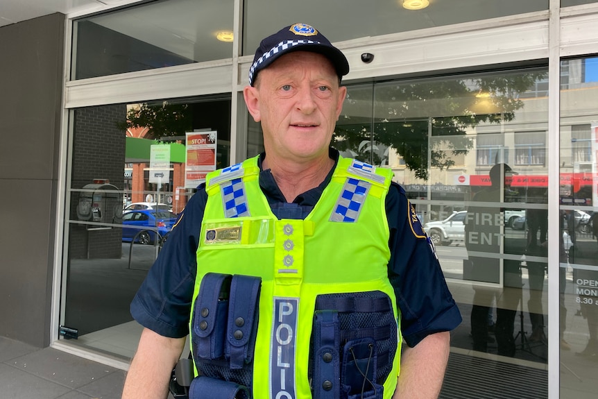 A police officer in a hi-vis vest and cap stands outside a building.