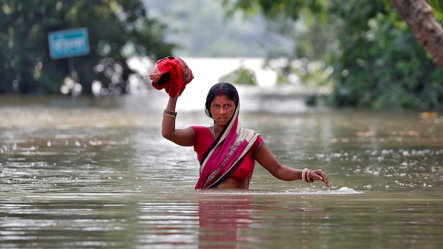 A woman wades through a flooded village while holding some cloth above her head.