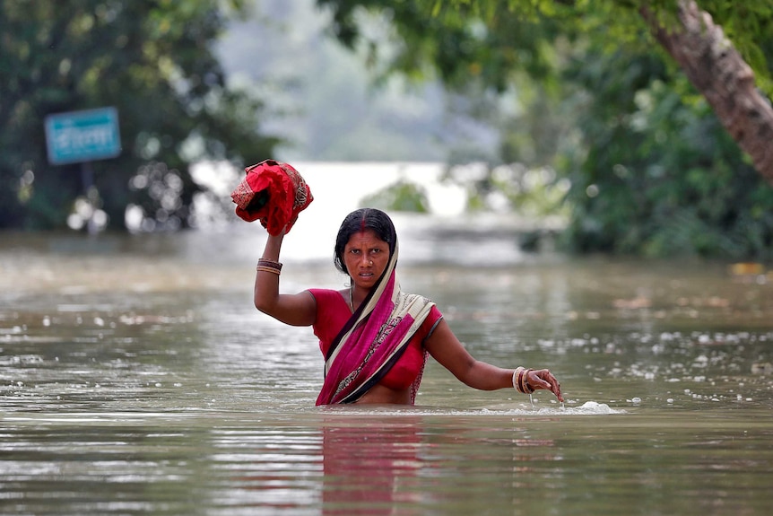 A woman wades through a flooded village while holding some cloth above her head.