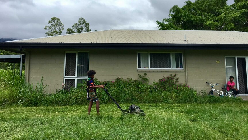 A young boy pushes a non-motorised lawn mower while his sister watches on near her bike.
