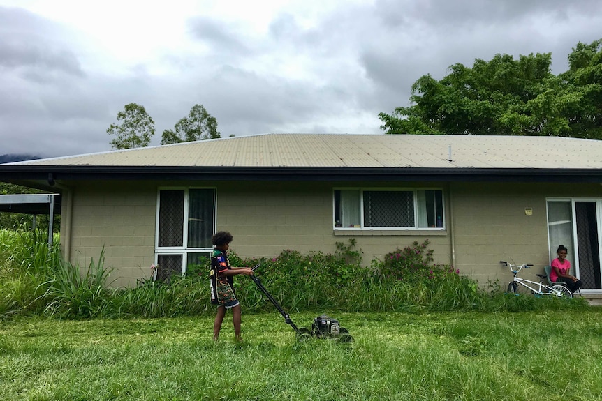 A young boy pushes a non-motorised lawn mower while his sister watches on near her bike.