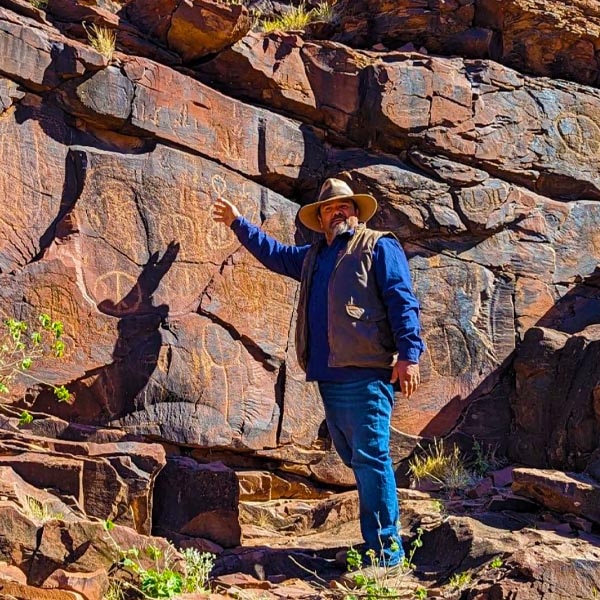 Kristian Coulthard in an Akubra-style hat. He points at red rocks carved with markings.