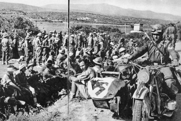 Australian troops sit on the ground while being guarded by a German on a motorcycle
