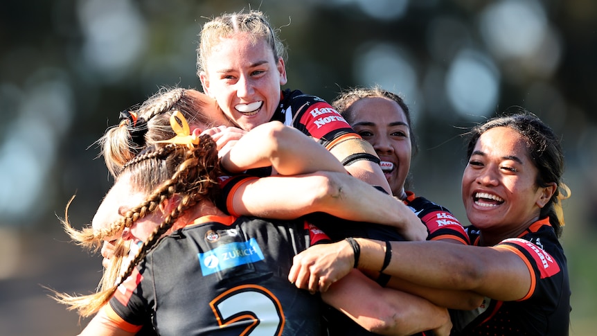 A Wests Tigers NRLW star grins in triumph as she jumps on top of a pile of teammates celebrating.