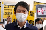 A man in a dark blue shirt and white face mask surrounded by protesters holding yellow signs