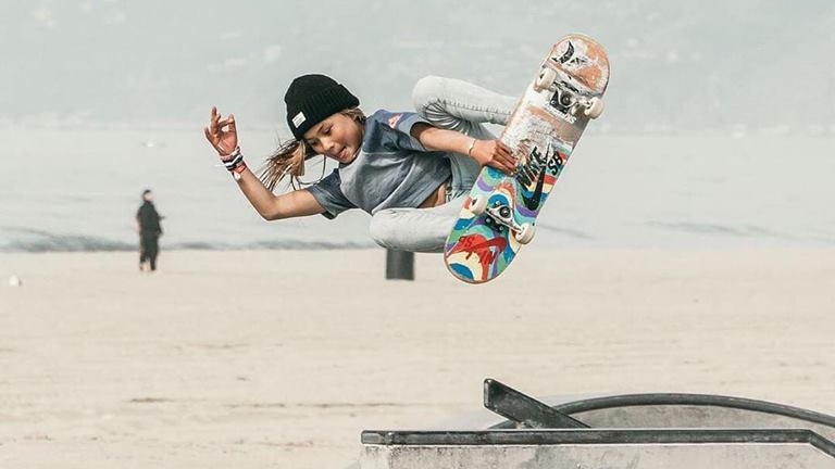 Sky Brown in the air holding a skateboard.