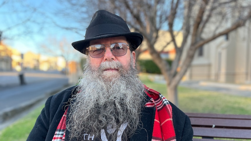 Man with long beard, black hat, red scarf sitting on a park bench