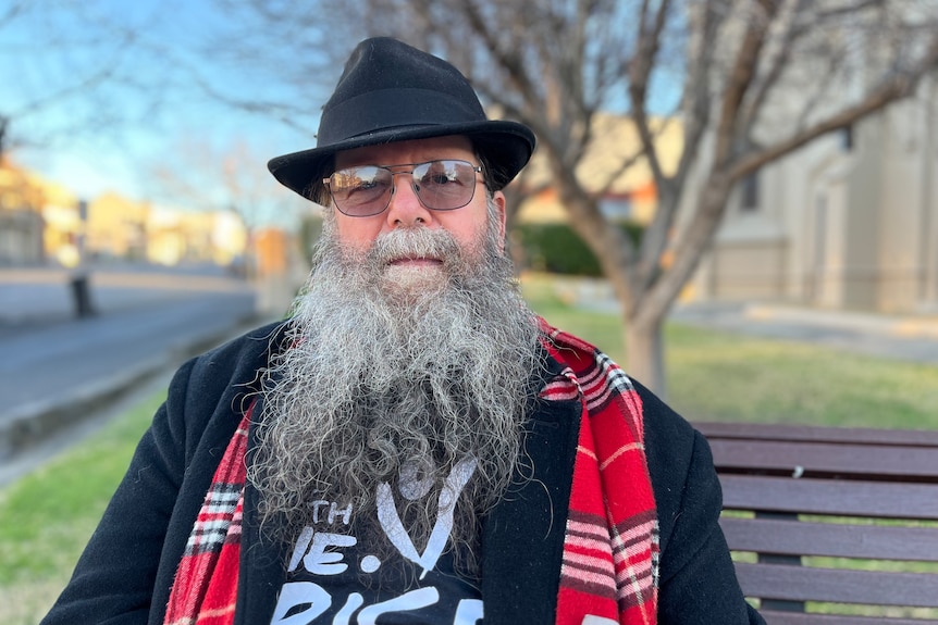 Man with long beard, black hat, red scarf sitting on a park bench