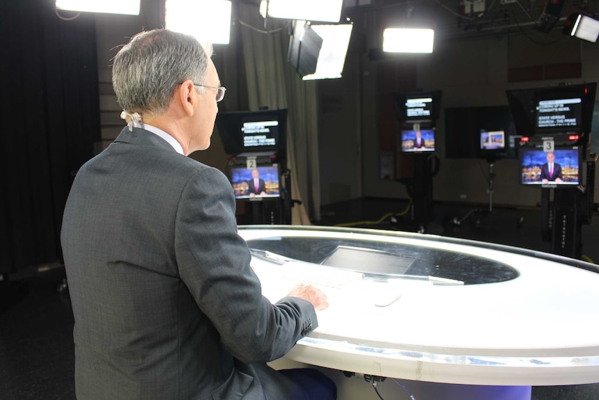 Rear shot of Gee at news desk in studio looking at camera and autocue.