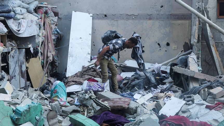 A man stands among the rubble of a house, looking down