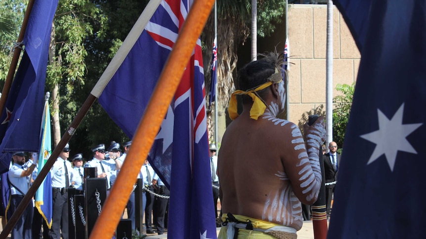 Kalkadoon man Ronaldo Gavara stands in traditional dress with a didgeridoo by his side, surrounded by Australian flags.