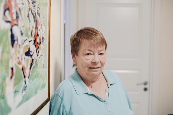 An older woman with short hear wearing a green shirt stands in front of a painting