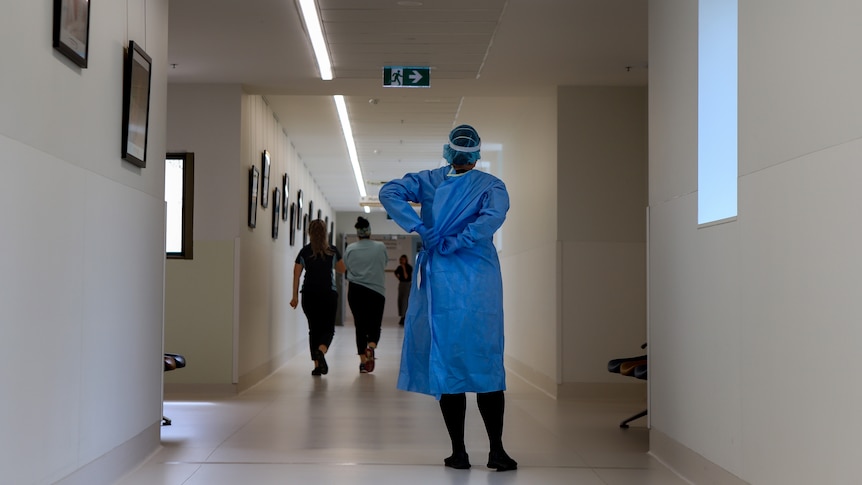 A nurse wearing adjust the back of her blue gown in a hospital corridor with people walking toward the end of the corridor