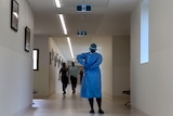 A nurse wearing adjust the back of her blue gown in a hospital corridor with people walking toward the end of the corridor