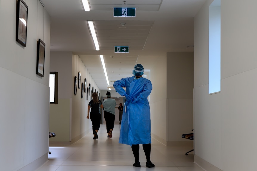 A nurse wearing adjust the back of her blue gown in a hospital corridor with people walking toward the end of the corridor