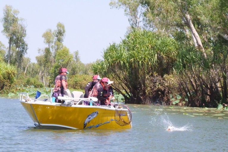 Four women stand on a yellow boat with a fishing net, with a fish splashing in the water