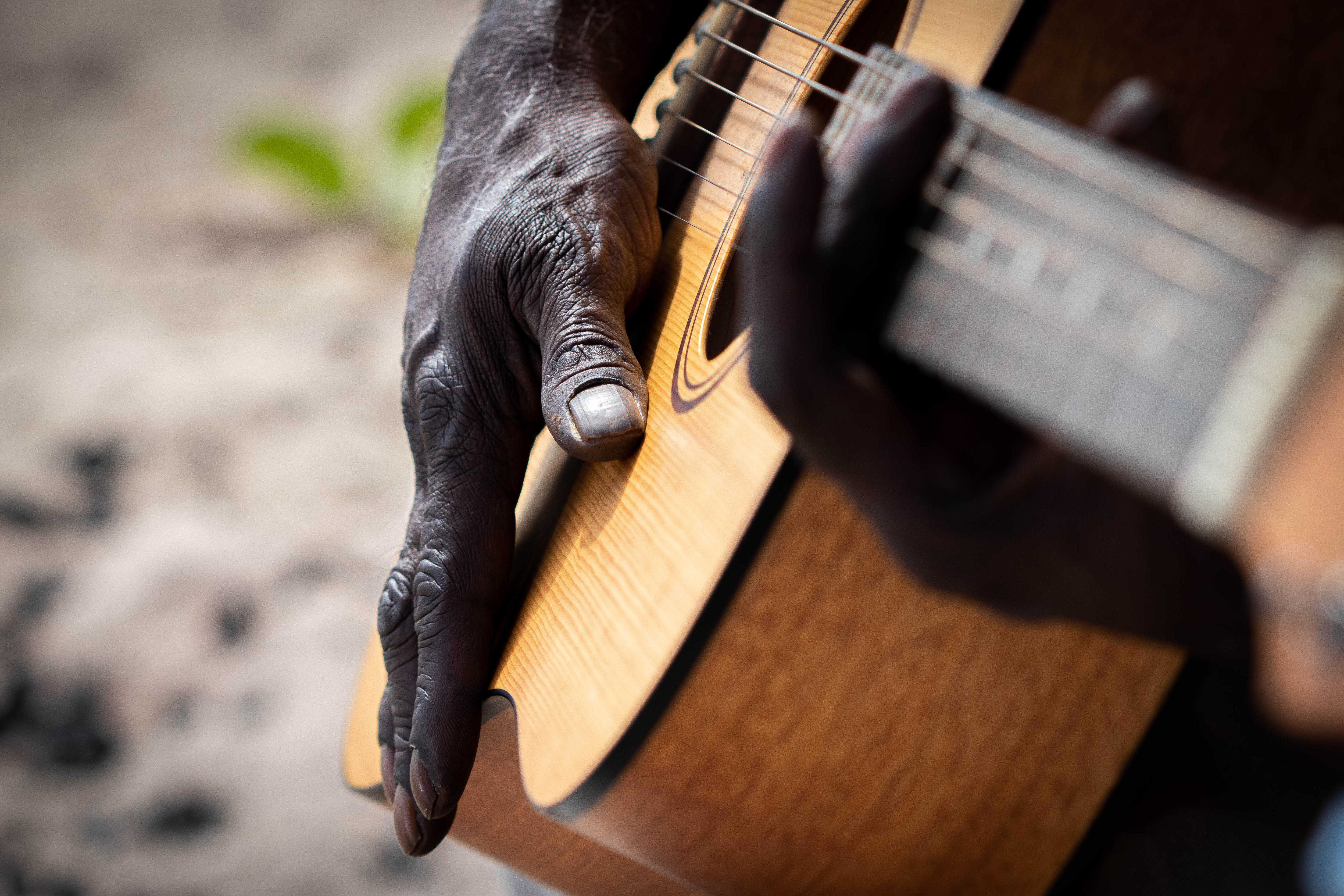 A man's hand plays a wooden guitar. The person is clearly outside, with green leaves and sand visible behind the instrument.