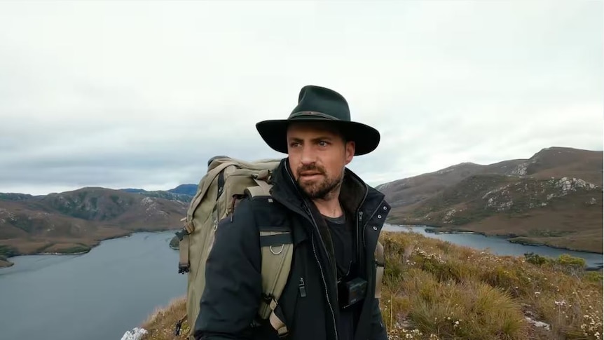 Portrait of man in landscape. He wears cowboy hat. Behind him is a lake and mountains.
