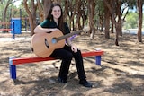 A girl holding a guitar sits on a bench in a school playground