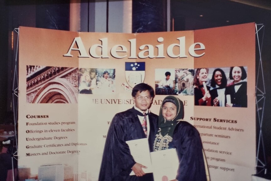 Woman in hijab stands in graduation gown next to man, both hold papers under arms. 